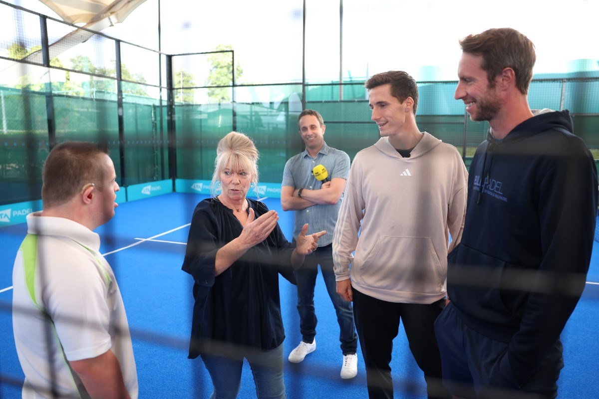 A woman stands in the middle of a group of men, making hand gestures towards one of the group. The two men oin the right of ther group are wearing hoodies, while the man in the middle, stood in the background, is holding a microphone.